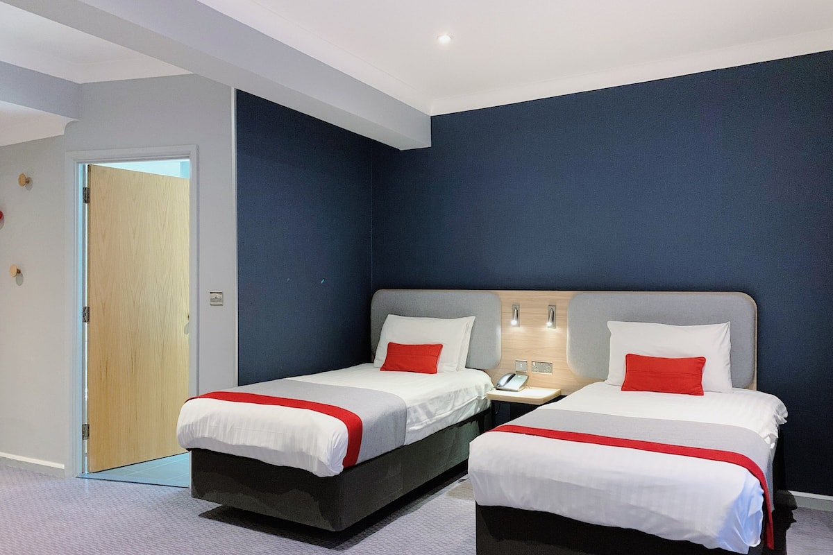 Quarto de hotel com duas camas, cada uma com roupa de cama branca e detalhes vermelhos, uma porta de madeira e uma parede em tom azul escuro.