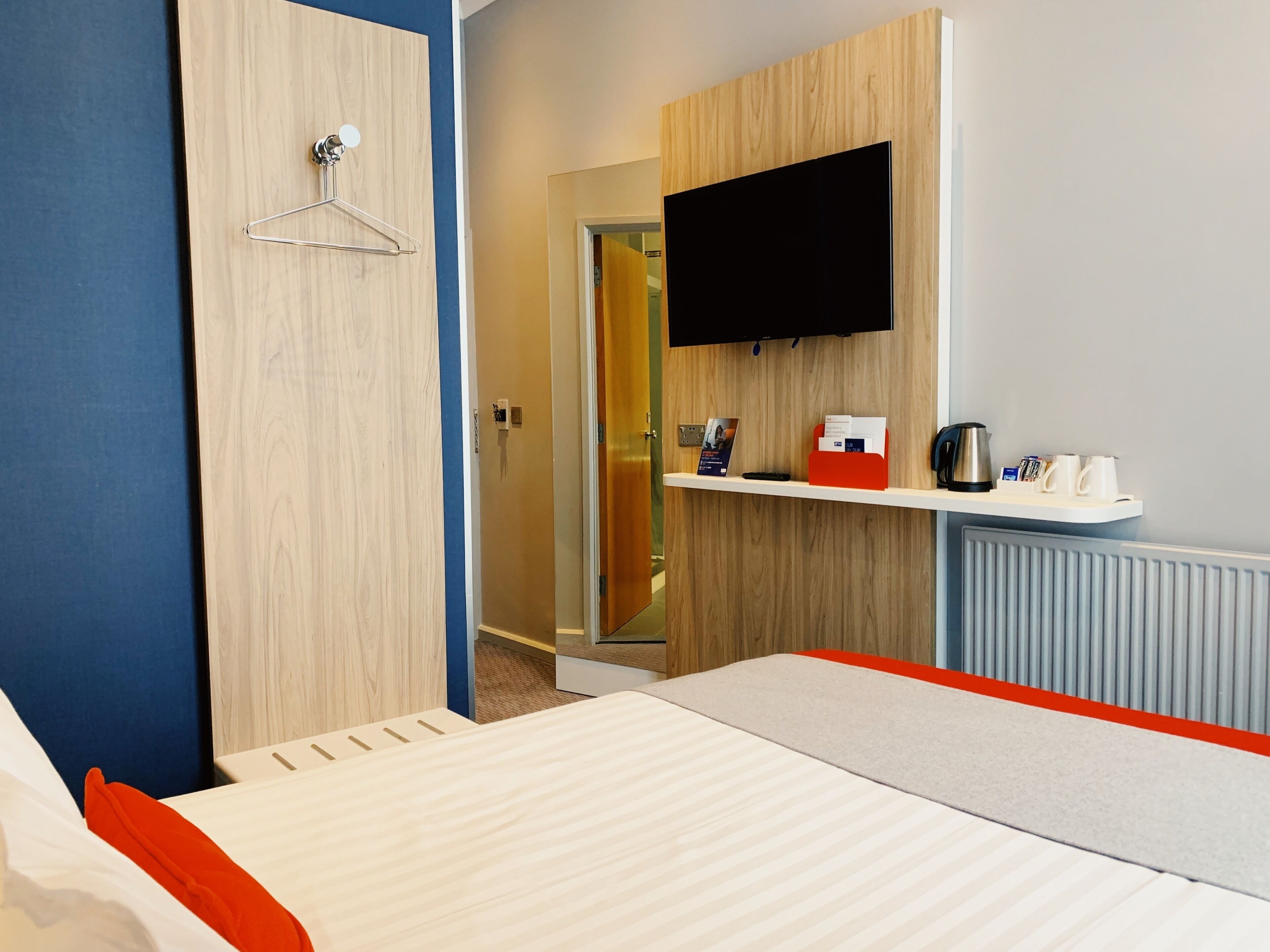 A hotel room with a bed, a TV mounted on the wall, a small shelf with a kettle and cups, and a wooden wardrobe.