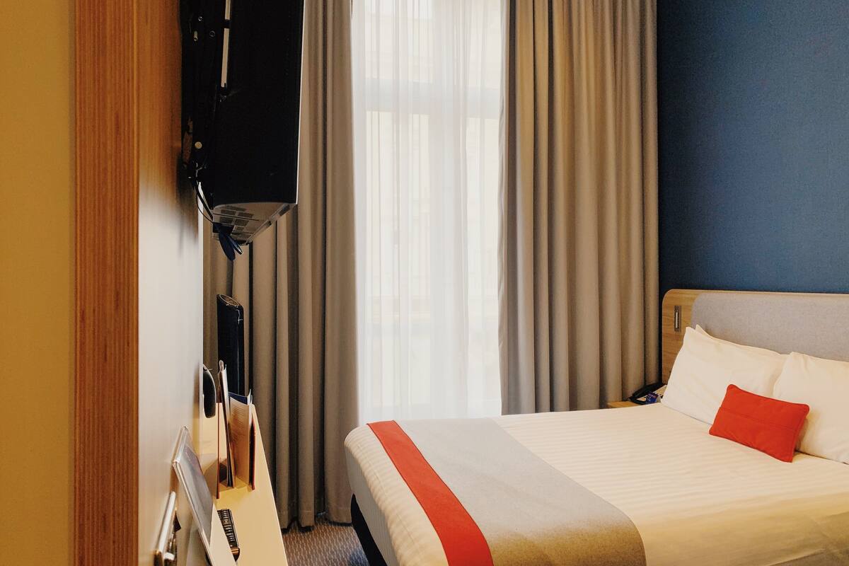 Una habitación de hotel con cama, escritorio con lámpara y televisor en la pared.