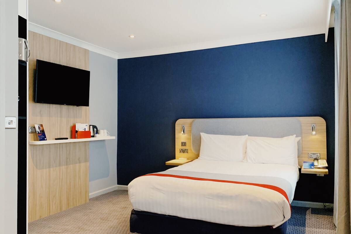 Habitación de hotel con una cama grande, un televisor de pantalla plana montado en la pared, una mesita con un teléfono y un libro, y una estantería con una tetera y tazas.