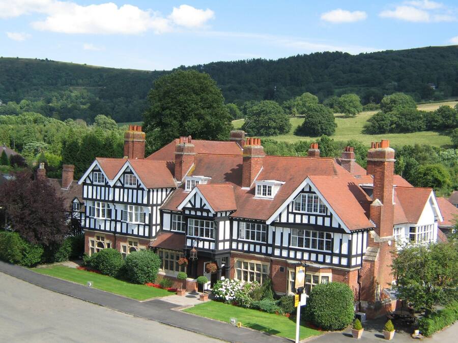 A traditional Tudor-style building with multiple chimneys, surrounded by lush greenery and hills.