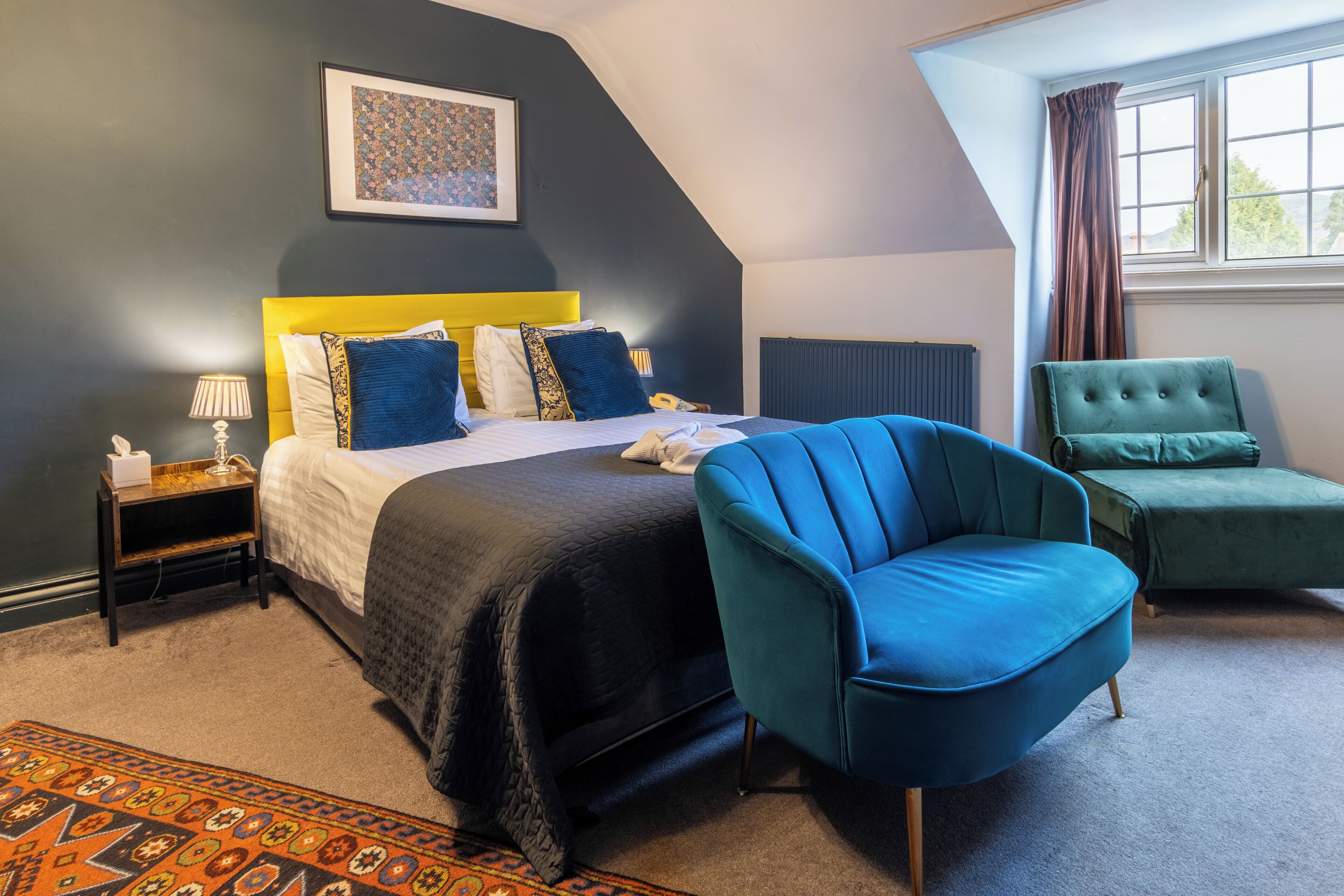 A bedroom with a bed, a blue armchair, a green armchair, a nightstand, a lamp, and a patterned rug.