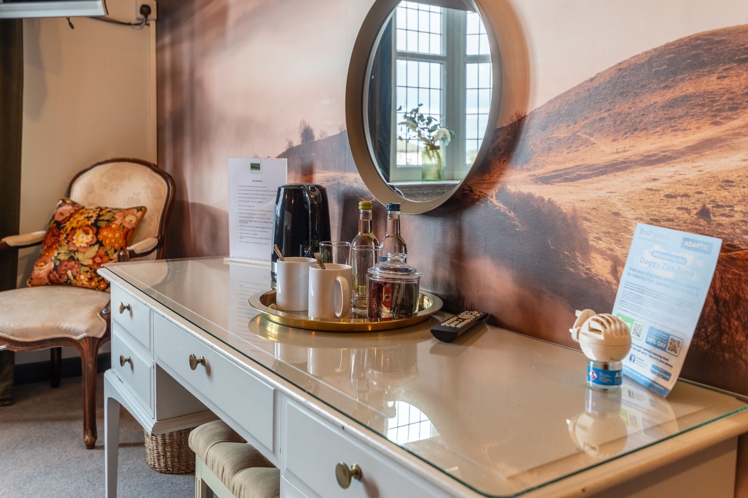 Ein Hotelzimmer mit Glastisch, einem Stuhl mit Blumekissen, einem Tablett mit Getränken und einer Fernbedienung, sowie einem Spiegel, der ein Fenster reflektiert.