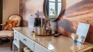 Ein Hotelzimmer mit Glastisch, einem Stuhl mit Blumekissen, einem Tablett mit Getränken und einer Fernbedienung, sowie einem Spiegel, der ein Fenster reflektiert.
