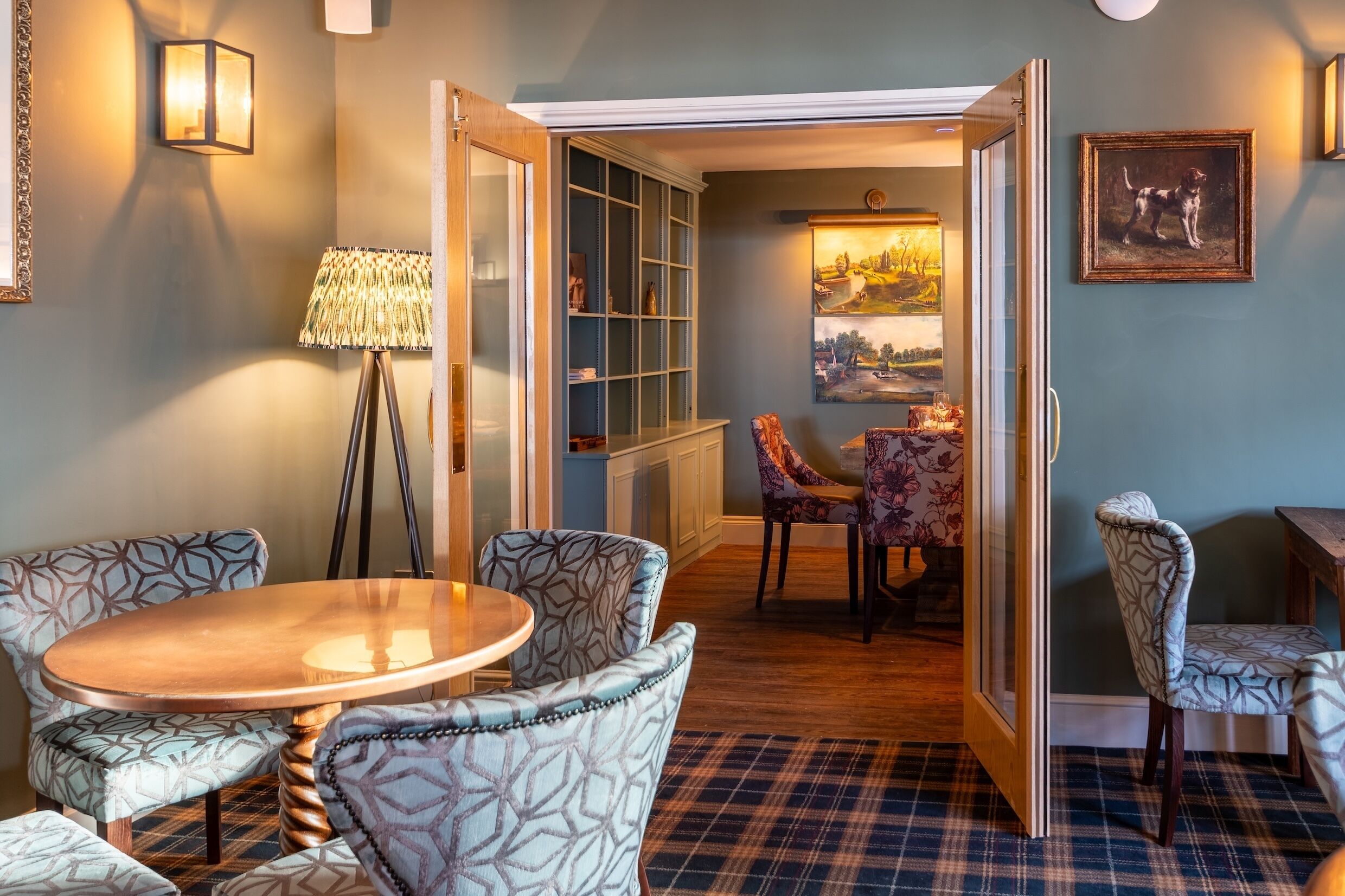 A room with patterned chairs, a round table, a lamp, a cabinet, and a painting on the wall.