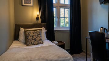 A bedroom with a bed, a bedside table with a lamp, a desk with a chair, and a window with curtains.