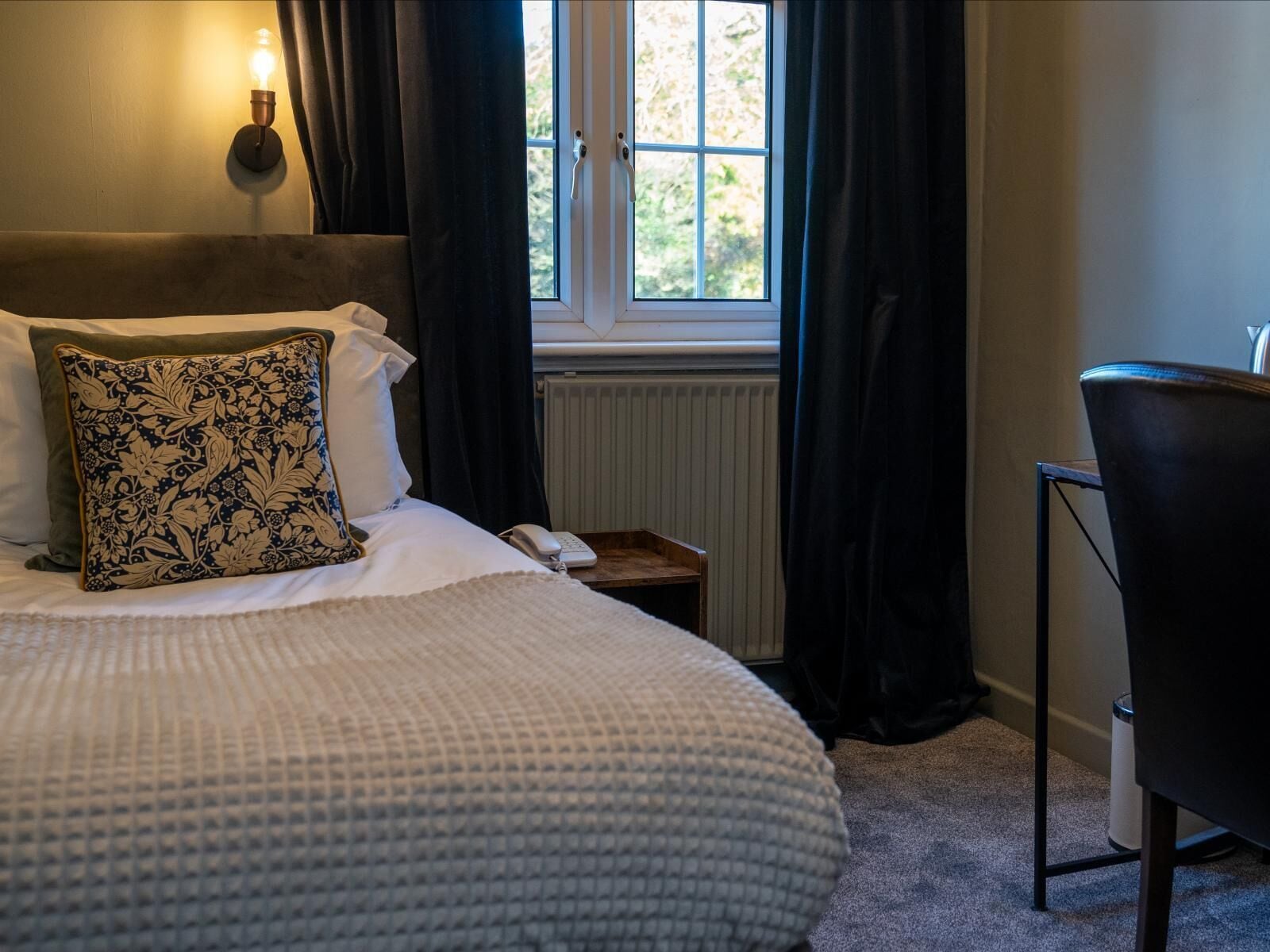 A neatly made bed with a patterned pillow, a bedside table with a phone, and a chair.