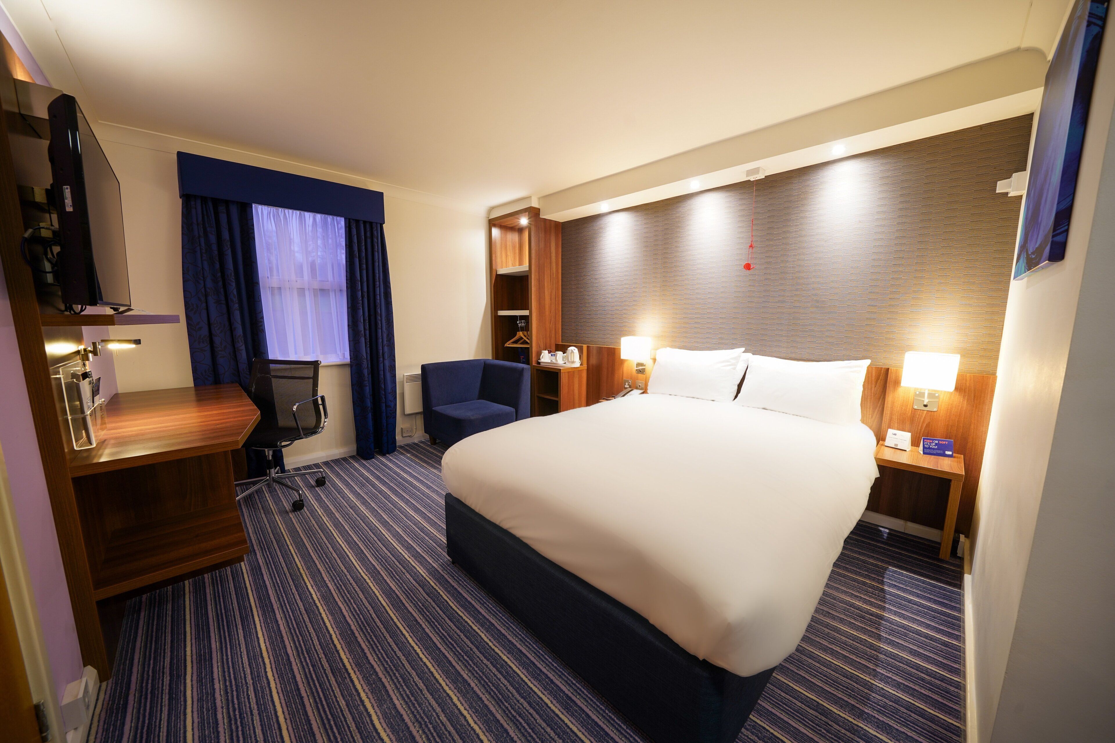 A hotel room with a large bed, a desk, a chair, a TV, and a small table with a lamp.