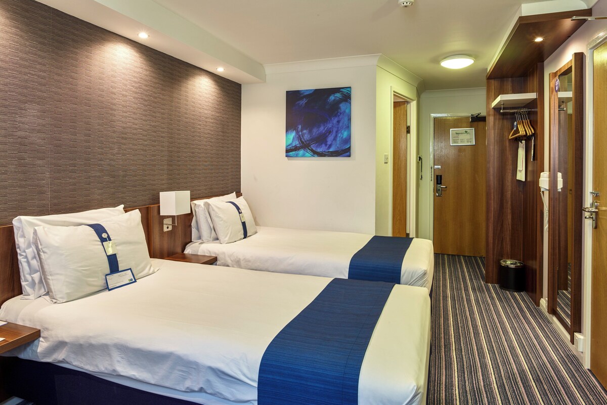 A hotel room with two beds, a wooden headboard, a painting on the wall, and a hallway with a coat rack.