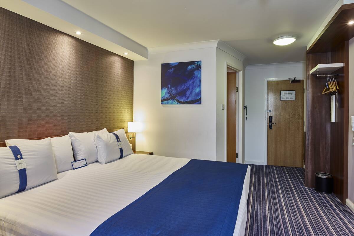 A hotel room with a large bed, a wooden headboard, a wall-mounted painting, a door with a sign, and a coat rack.