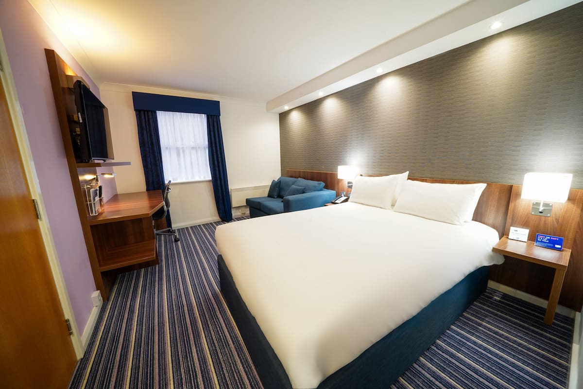 A hotel room with a large bed, a desk, a chair, a TV, and a window with curtains.