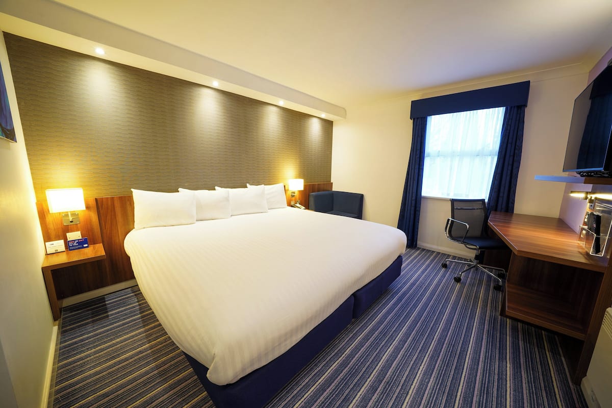 A hotel room with a large bed, a desk, a chair, a TV, and a window with curtains.