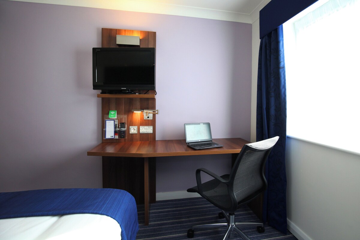 A hotel room with a bed, a desk with a laptop, a TV, and a window with curtains.