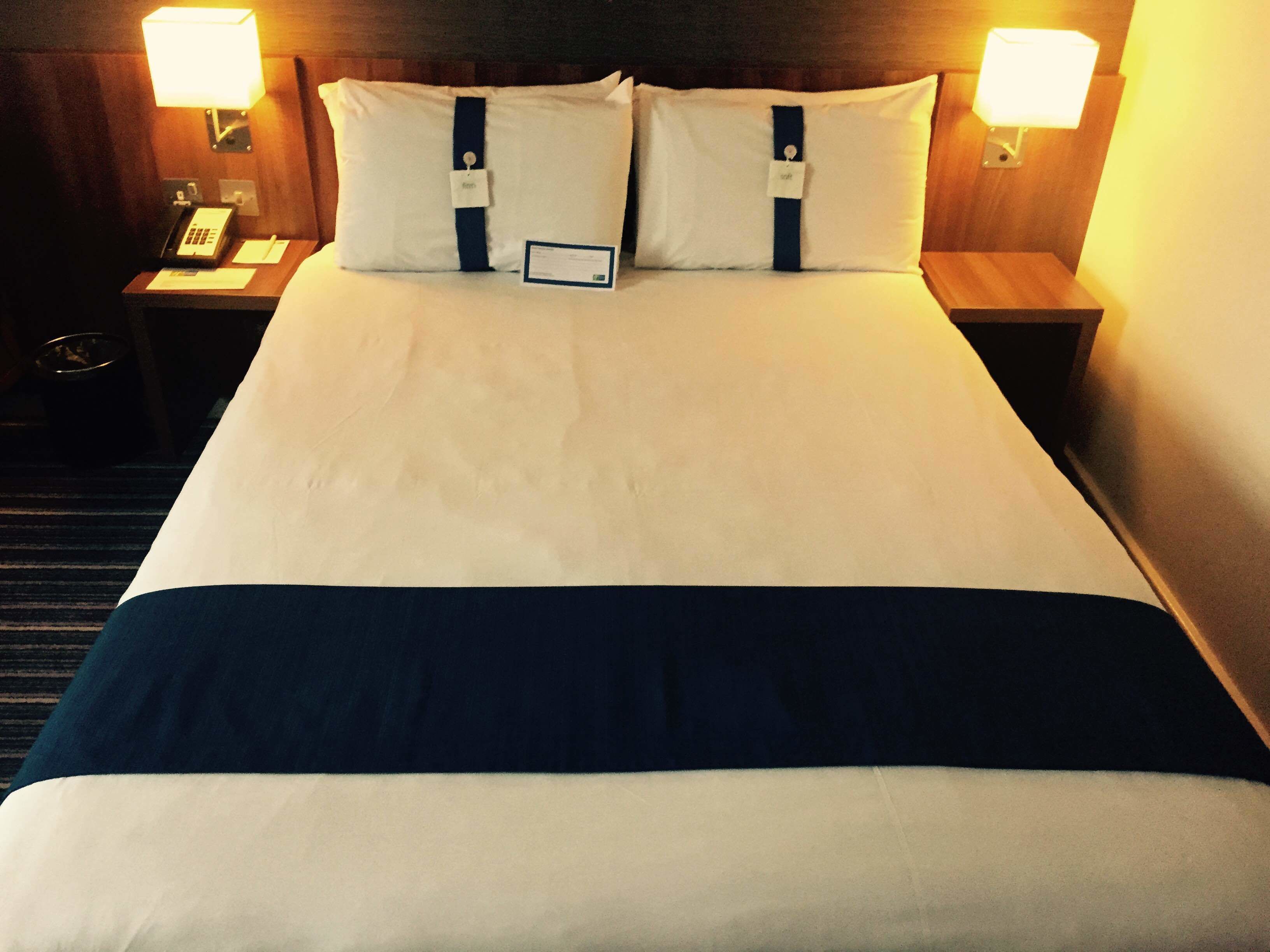 A hotel room with a neatly made bed, two bedside lamps, a nightstand with a telephone, and a sign indicating the room number.