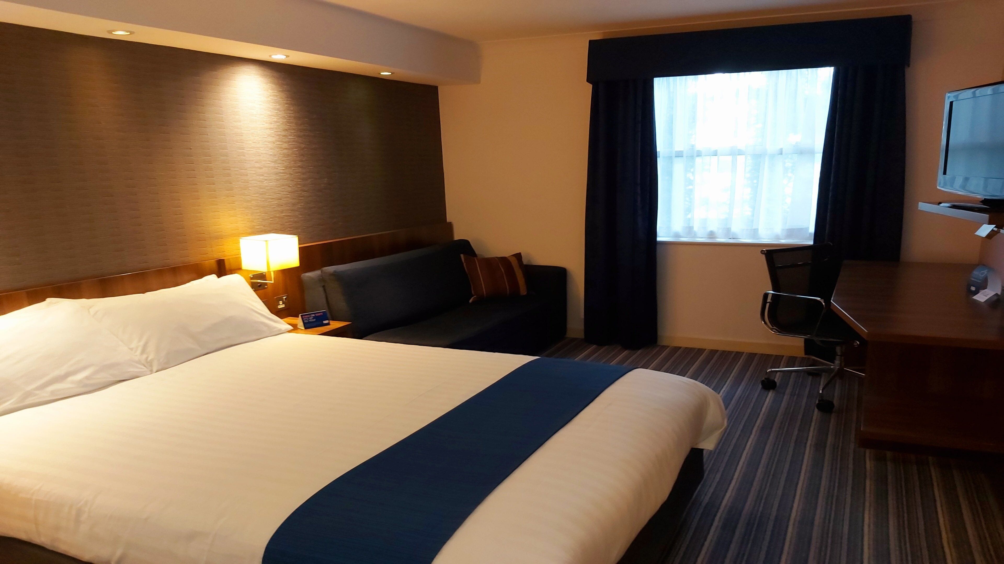 A hotel room with a large bed, a desk, a chair, a TV, and a window with curtains.