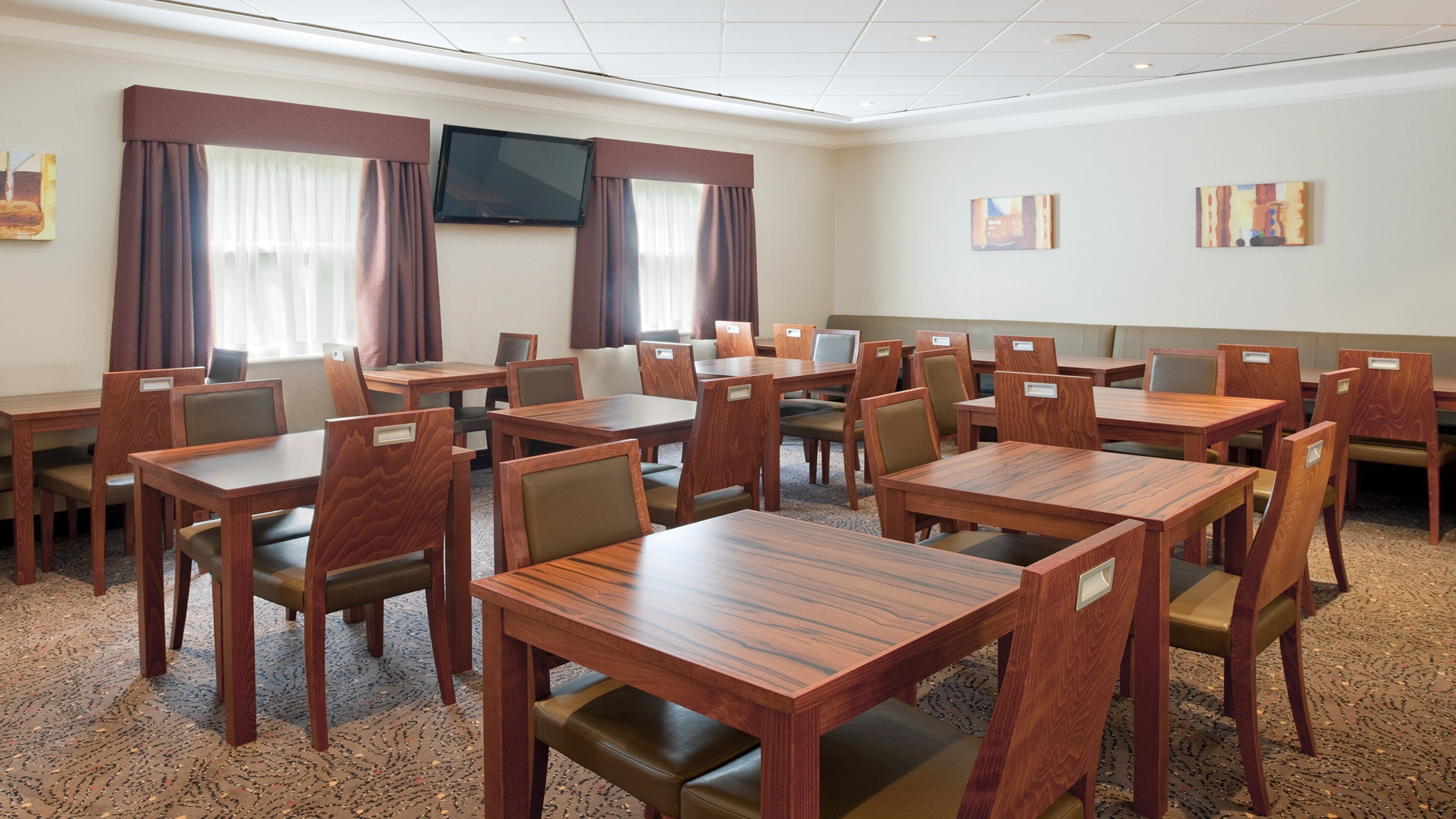 A hotel conference room with multiple tables and chairs, a flat-screen TV mounted on the wall, and artwork on the walls.