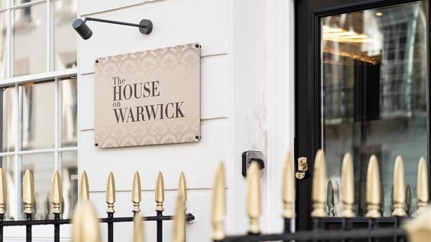En skylt med texten ”THE HOUSE ON WARWICK” är monterad på en vit vägg, och framför väggen finns ett svart metallstaket.