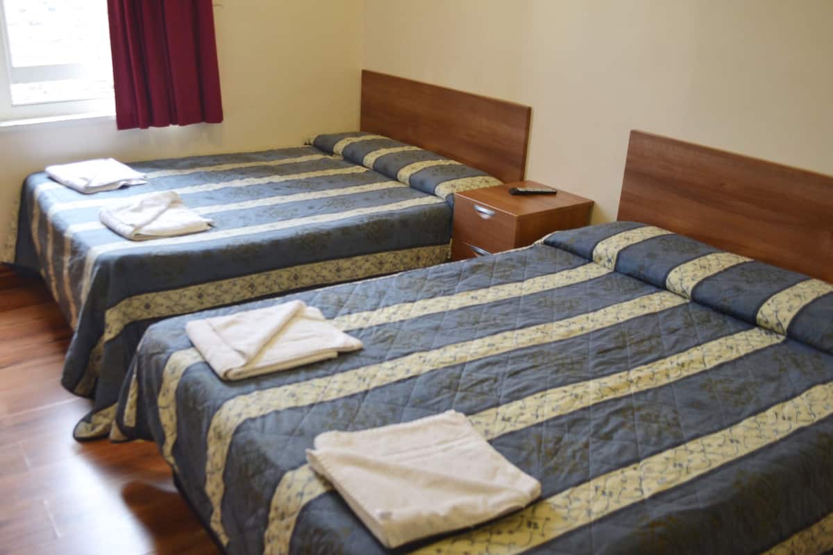 A hotel room with three single beds, each with a wooden headboard and a blue and yellow striped bedspread. There is a wooden nightstand between the beds with a remote control on it.