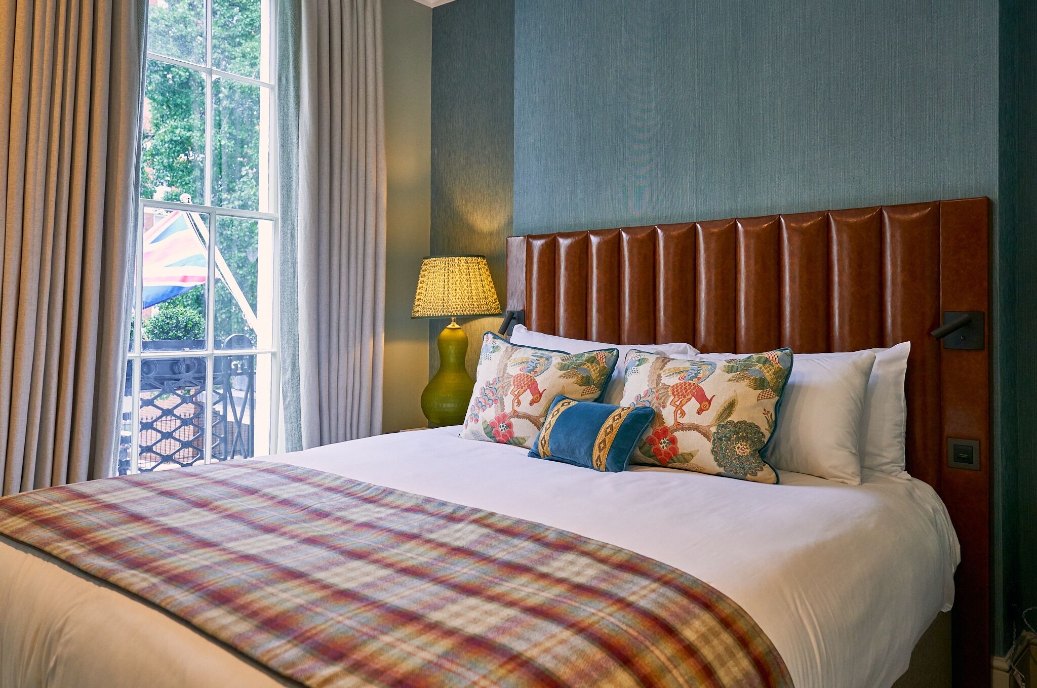 A hotel room with a large bed, a headboard, a window with curtains, and a lamp.