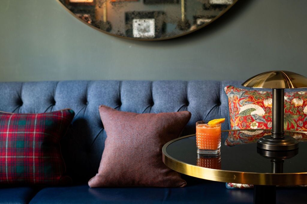 Ein blaues Sofa mit Kissen in Karomuster und einfarbigen Designs, ein runder Tisch mit einem Getränk und eine dekorative Lampe.