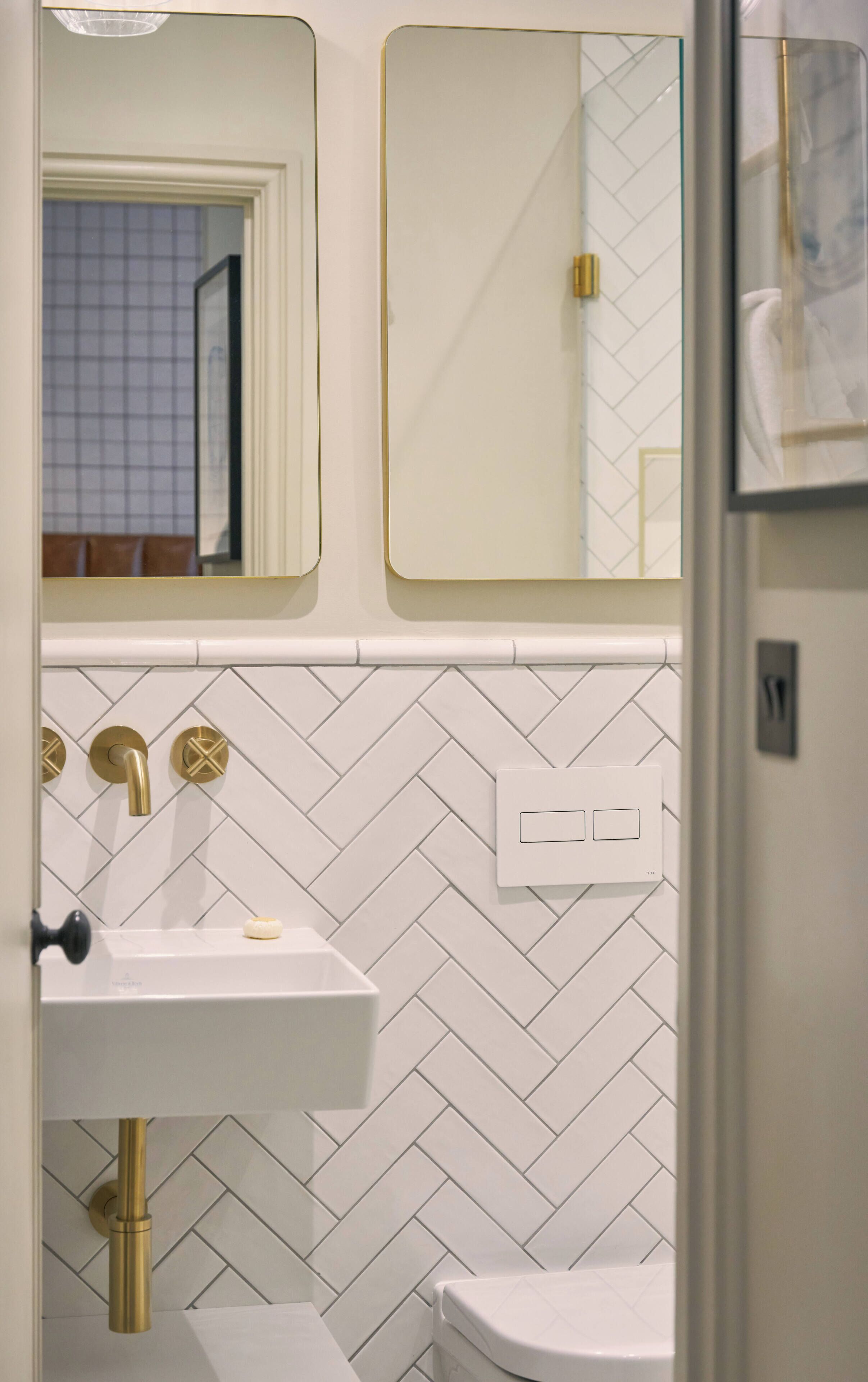 A bathroom with a white herringbone tile backsplash, a white sink with gold fixtures, and a toilet.
