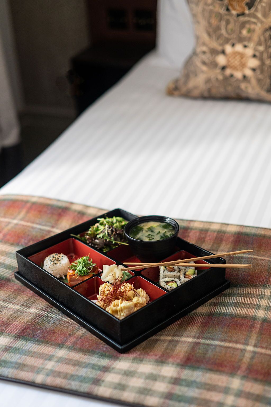 A meal served in a black bento box on a plaid tablecloth, with chopsticks and a pillow in the background.