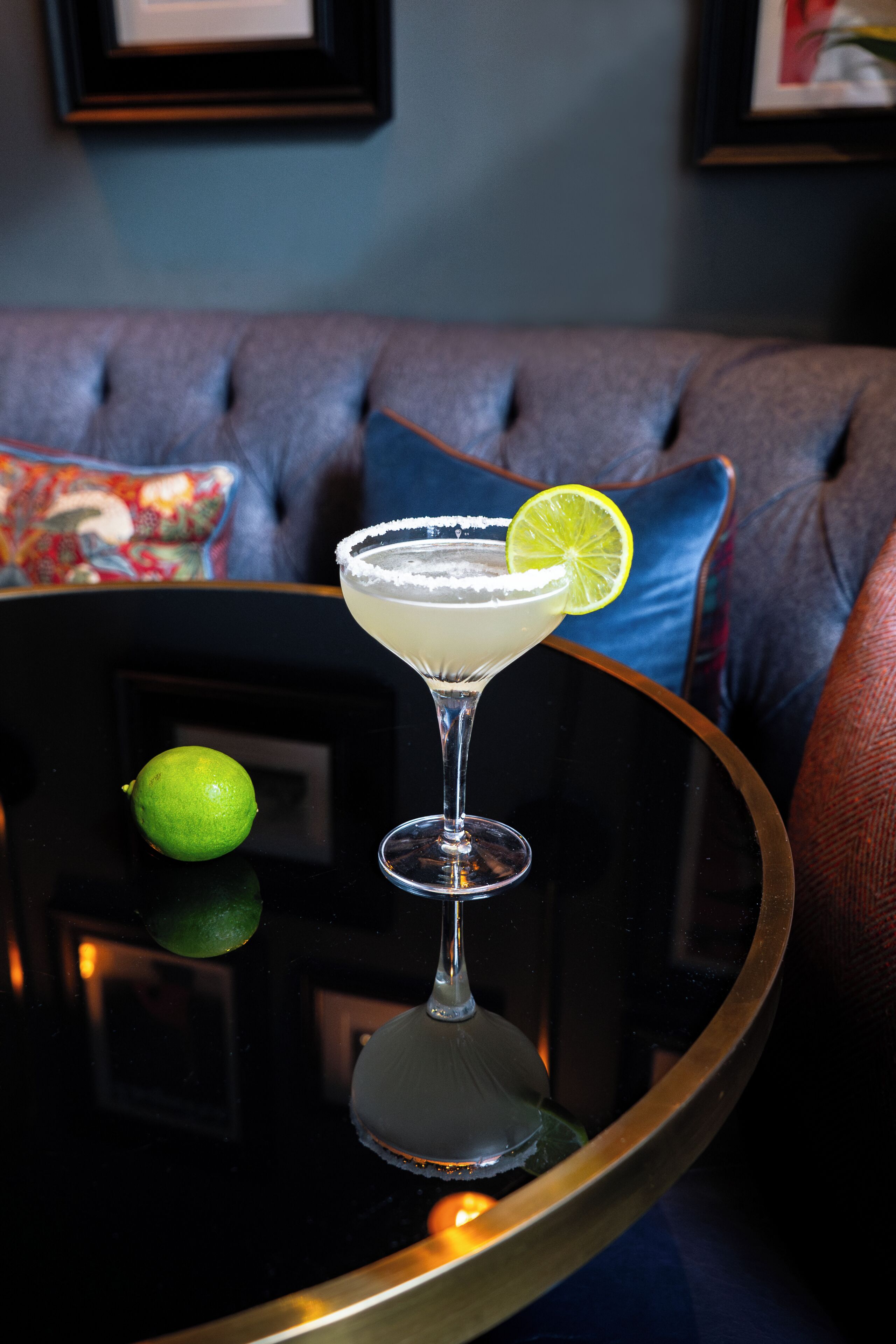 Un cocktail su un tavolo con guarnizione di lime, un lime e un divano sullo sfondo.