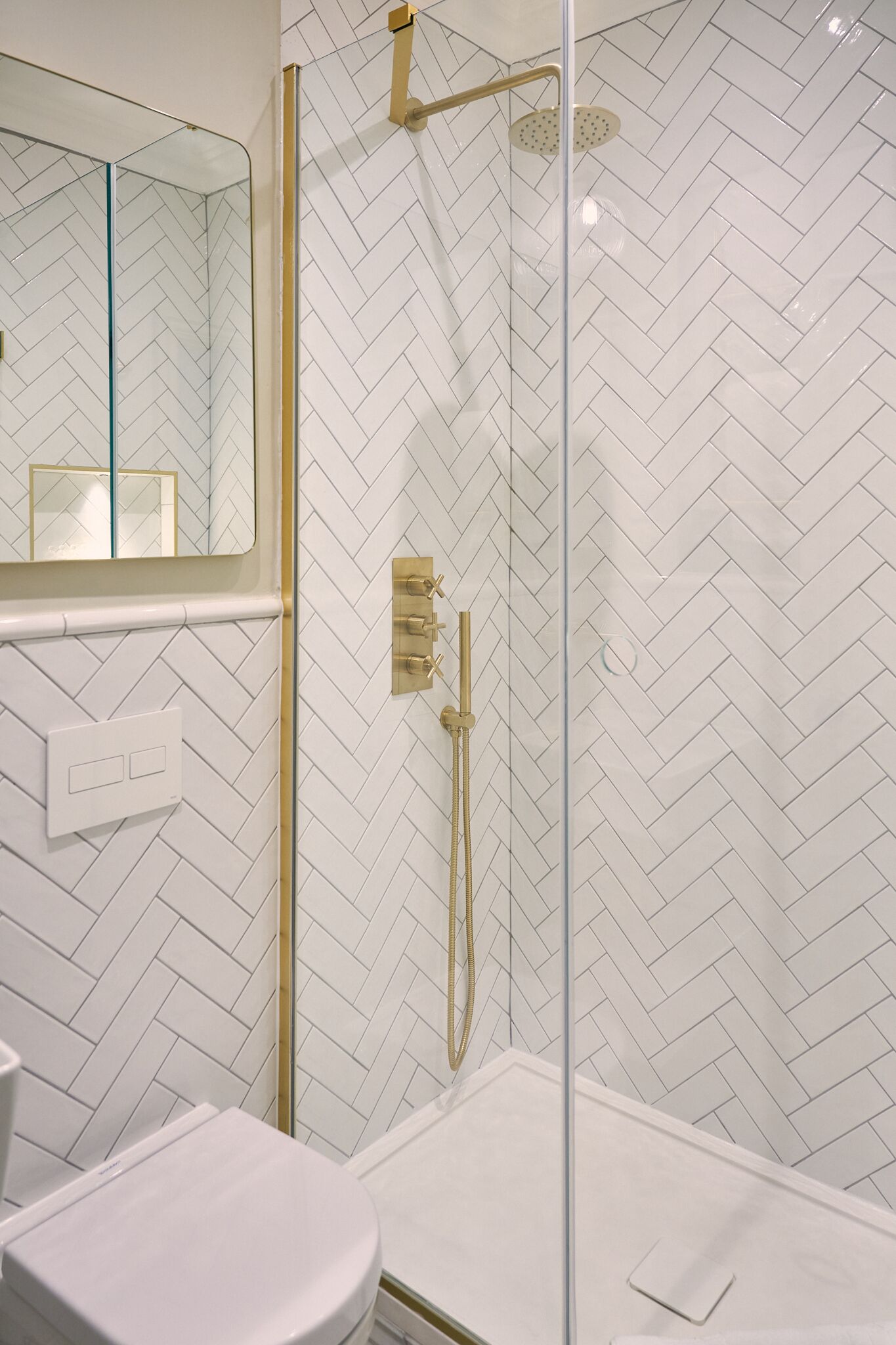 Une salle de bain moderne avec une douche à l’italienne en verre, des carreaux blancs disposés en motif chevrons, une pomme de douche dorée et des toilettes blanches.