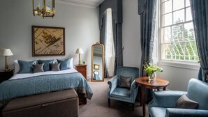 A hotel room with a bed, two armchairs, a round table, a mirror, a chandelier, and a window with curtains.