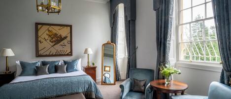 Hotelkamer met een bed, twee fauteuils, een ronde tafel, een spiegel, een kroonluchter en een raam met gordijnen.