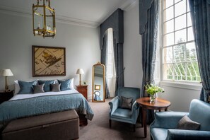 A hotel room with a bed, two armchairs, a round table, a mirror, a chandelier, and a window with curtains.