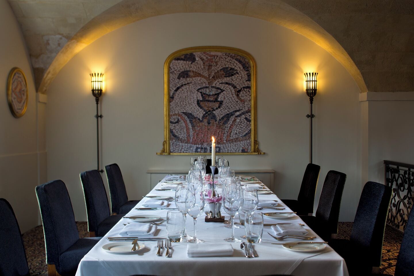 Une longue table à manger, dressée pour un repas, avec des chaises de chaque côté, un chandelier avec une bougie allumée et une œuvre d’art en mosaïque au-dessus.