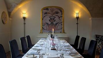 A long dining table set for a meal with chairs on both sides, a candelabra with a lit candle, and a mosaic artwork above.