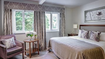 Un dormitorio con cama, una silla, una mesita con un florero y una ventana con cortinas.