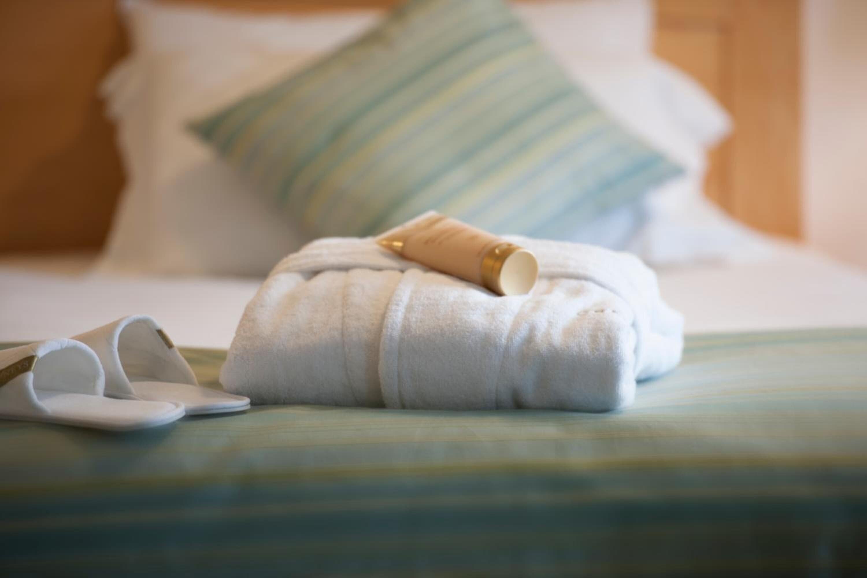 A neatly made bed with a striped blanket, a rolled towel, and a pair of slippers.