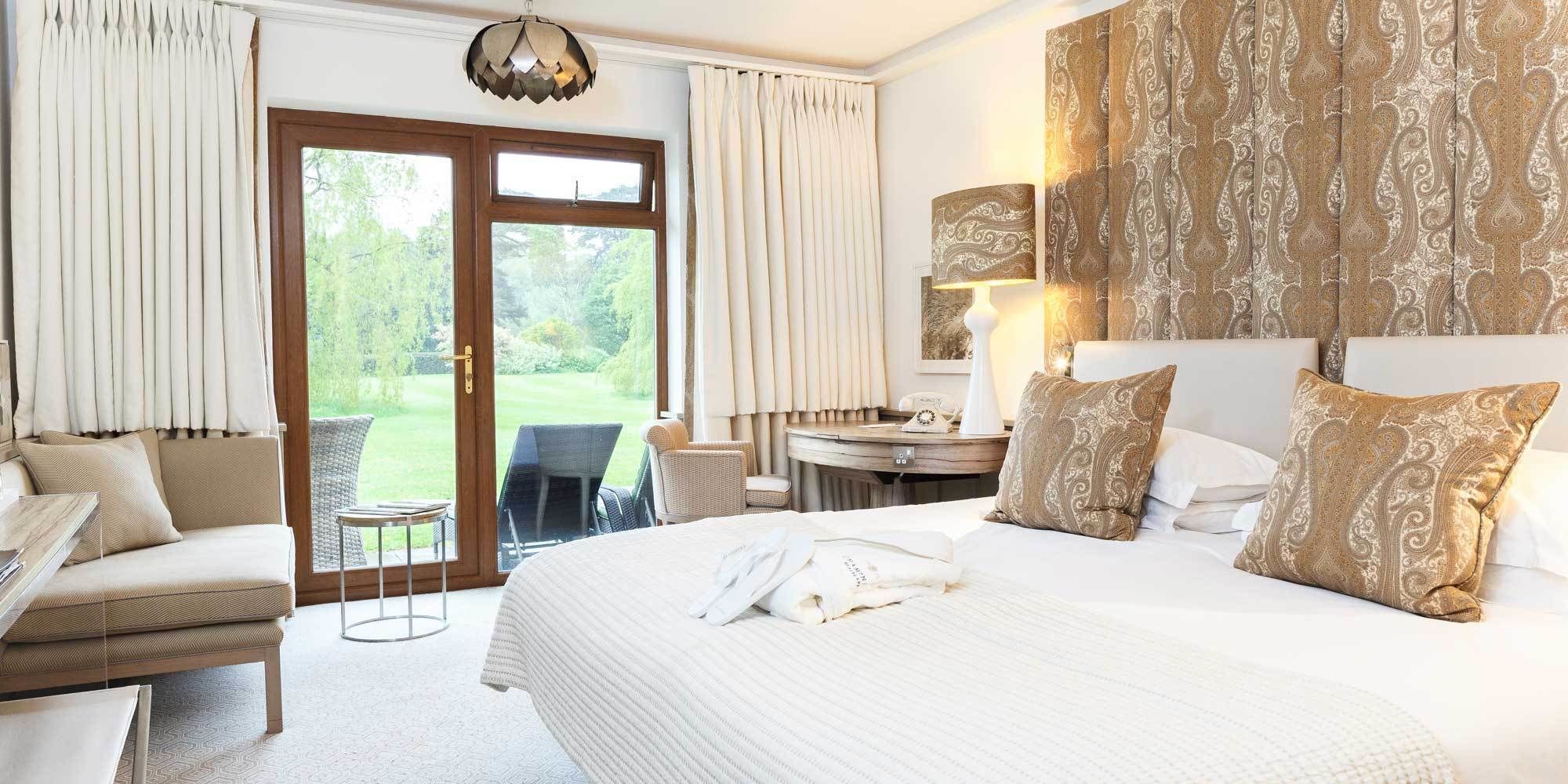 A hotel room with a large bed, a desk, a chair, and a view of a garden through the door.