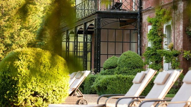 A sunlit outdoor area with lounge chairs, a covered patio, and a brick building with a balcony. - Champneys Tring (Tring)