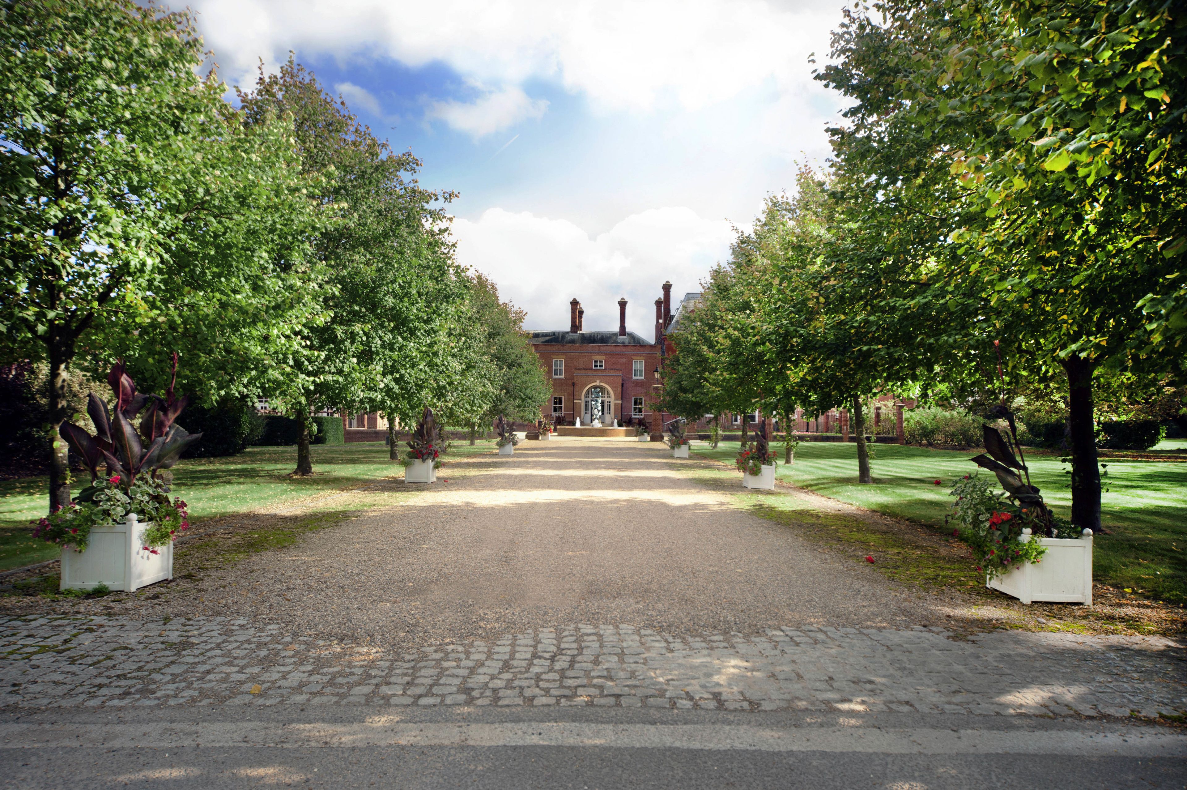 A cobblestone driveway leading to a brick building with a central entrance, flanked by symmetrical rows of trees and flower beds.