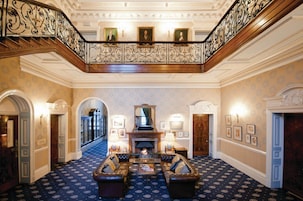 Um amplo hall com teto alto, grade de ferro ornamentada, lareira e um tapete estampado.