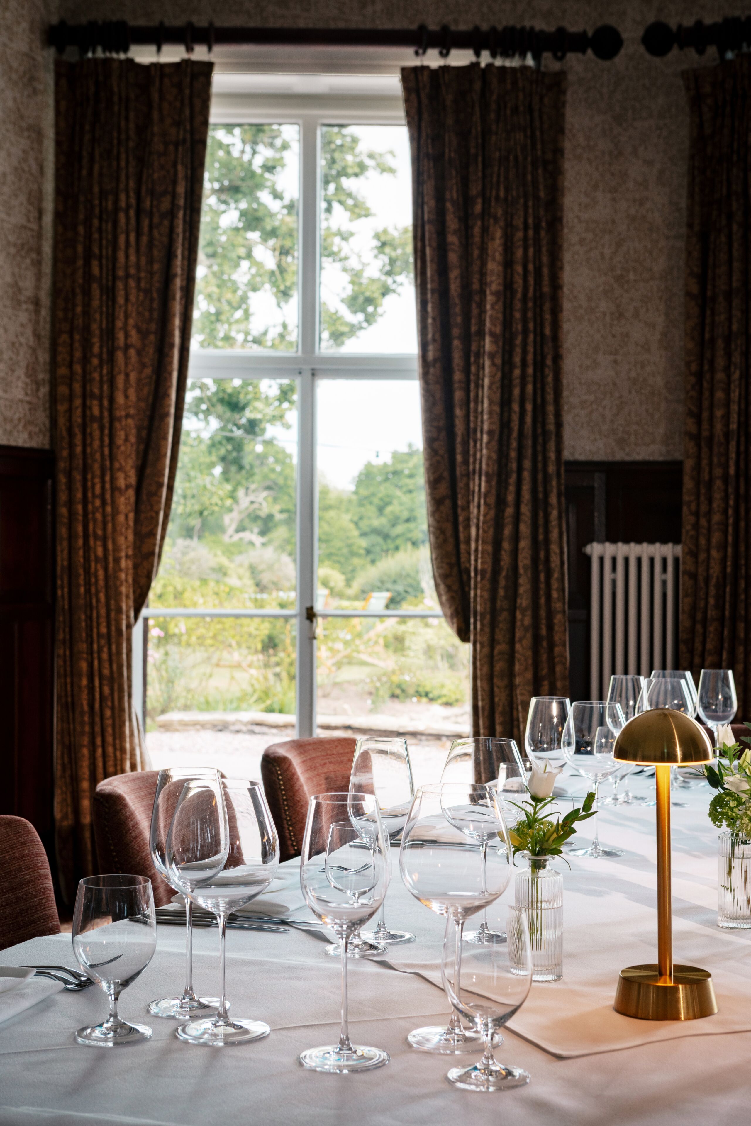 A dining table set with wine glasses and a gold lamp, with curtains and a view of greenery outside.