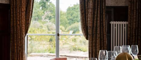 A dining table set with wine glasses and a gold lamp, with curtains and a view of greenery outside.