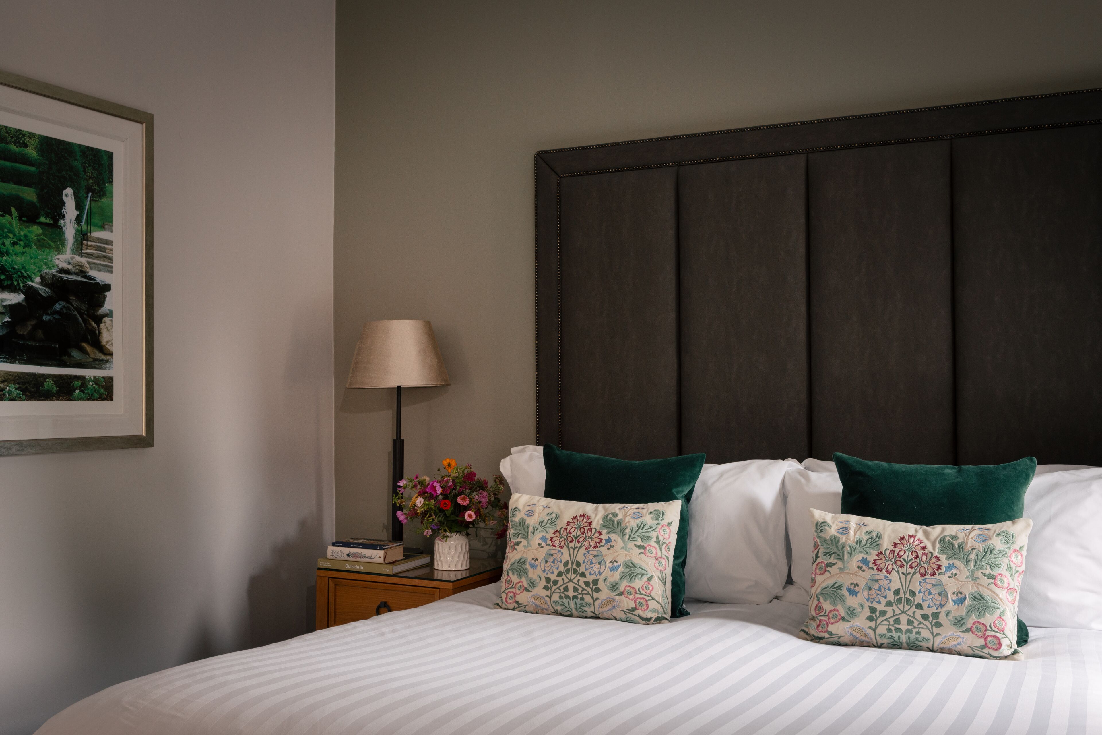 Uma cama bem arrumada com duas almofadas, uma mesinha de cabeceira com uma luminária e um vaso de flores, além de um quadro na parede.