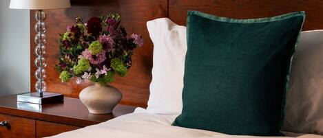 A neatly made bed with a white pillow and a green accent pillow, a bedside table with a lamp and a vase of flowers, and a wall-mounted abstract painting.