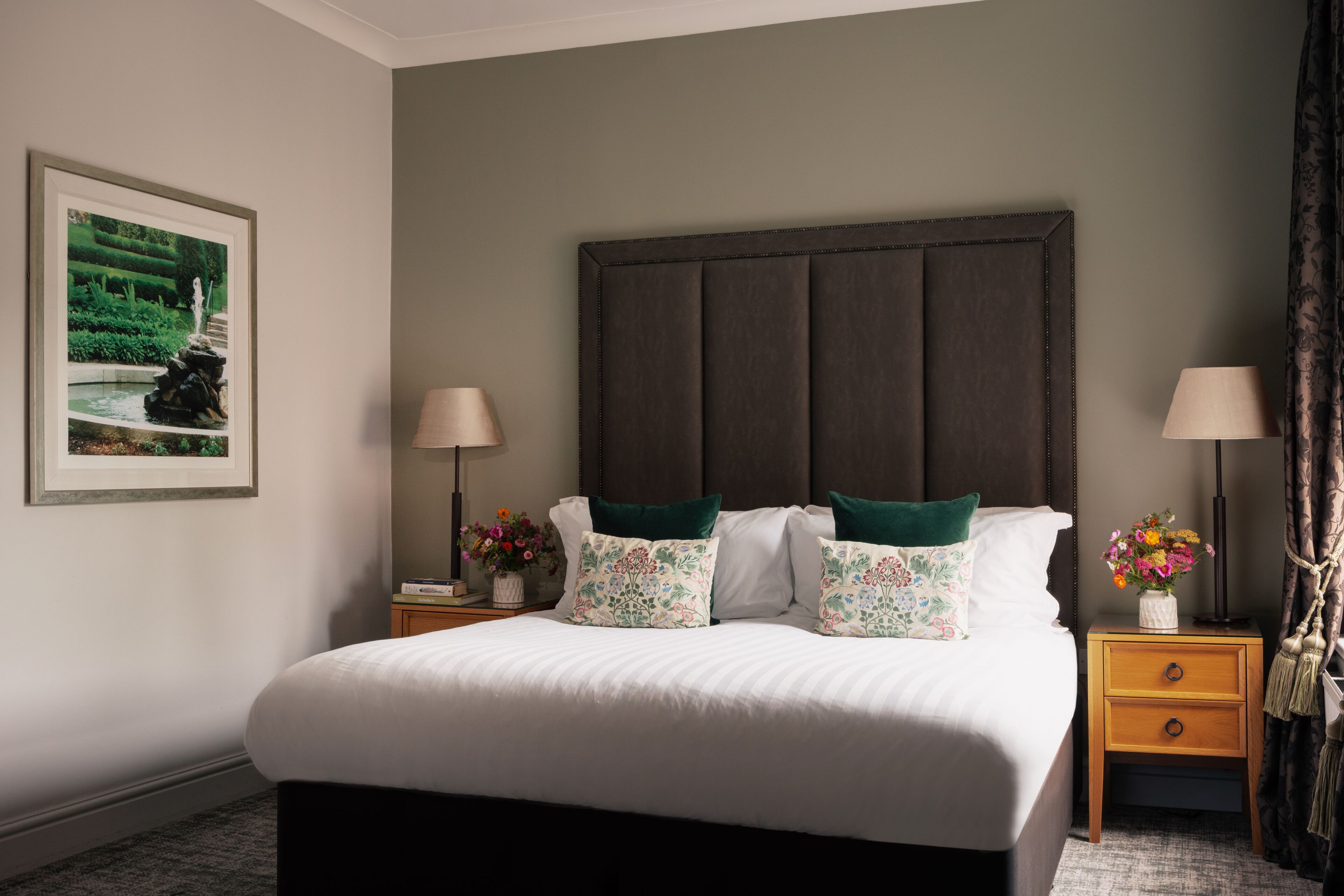 Een netjes opgemaakt bed met hoofdbord, twee nachtkastjes met lampen, een ingelijst kunstwerk aan de muur en een klein vaasje met bloemen op het nachtkastje.