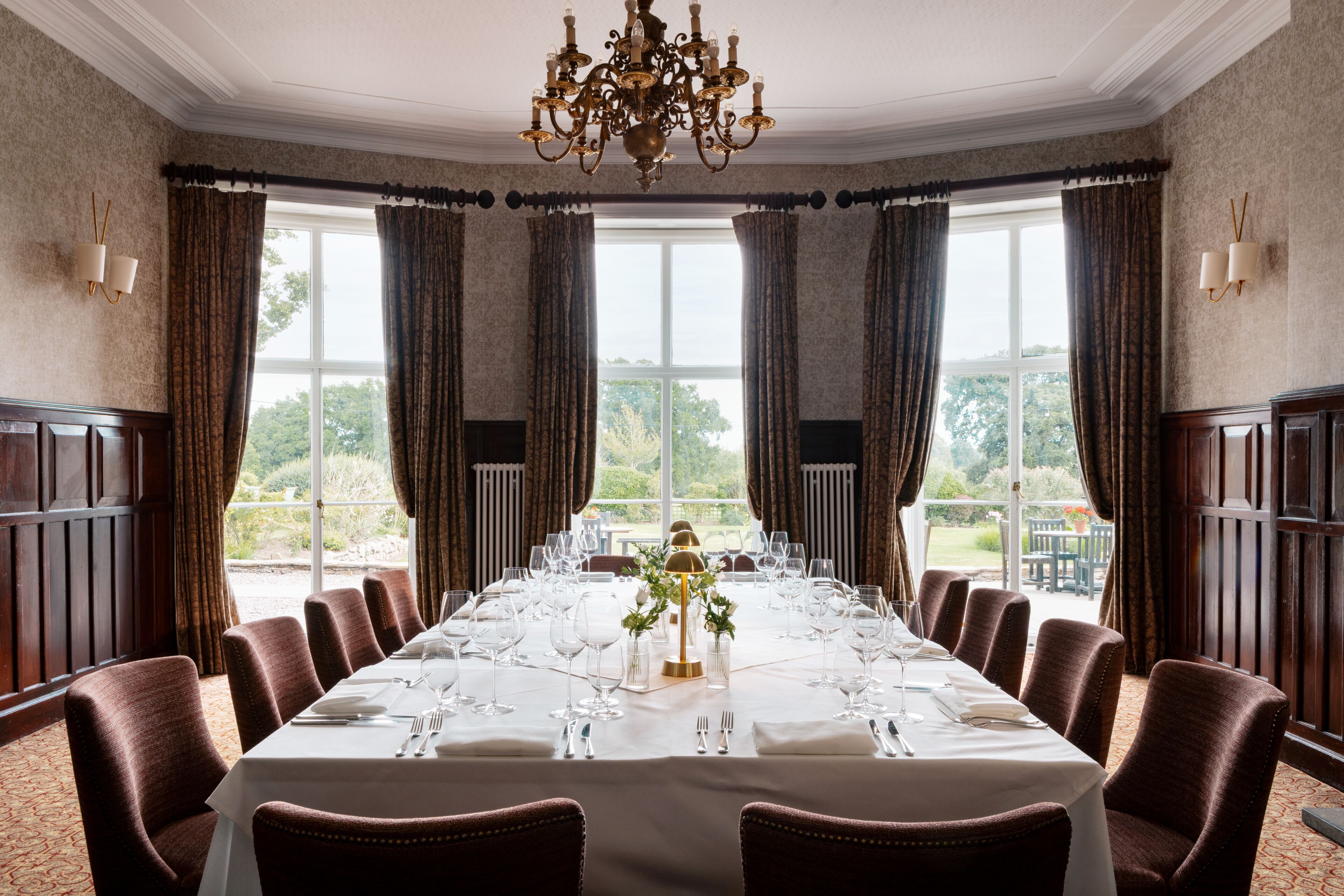 A formal dining room with a long table set for a meal, large windows with curtains, and a chandelier.