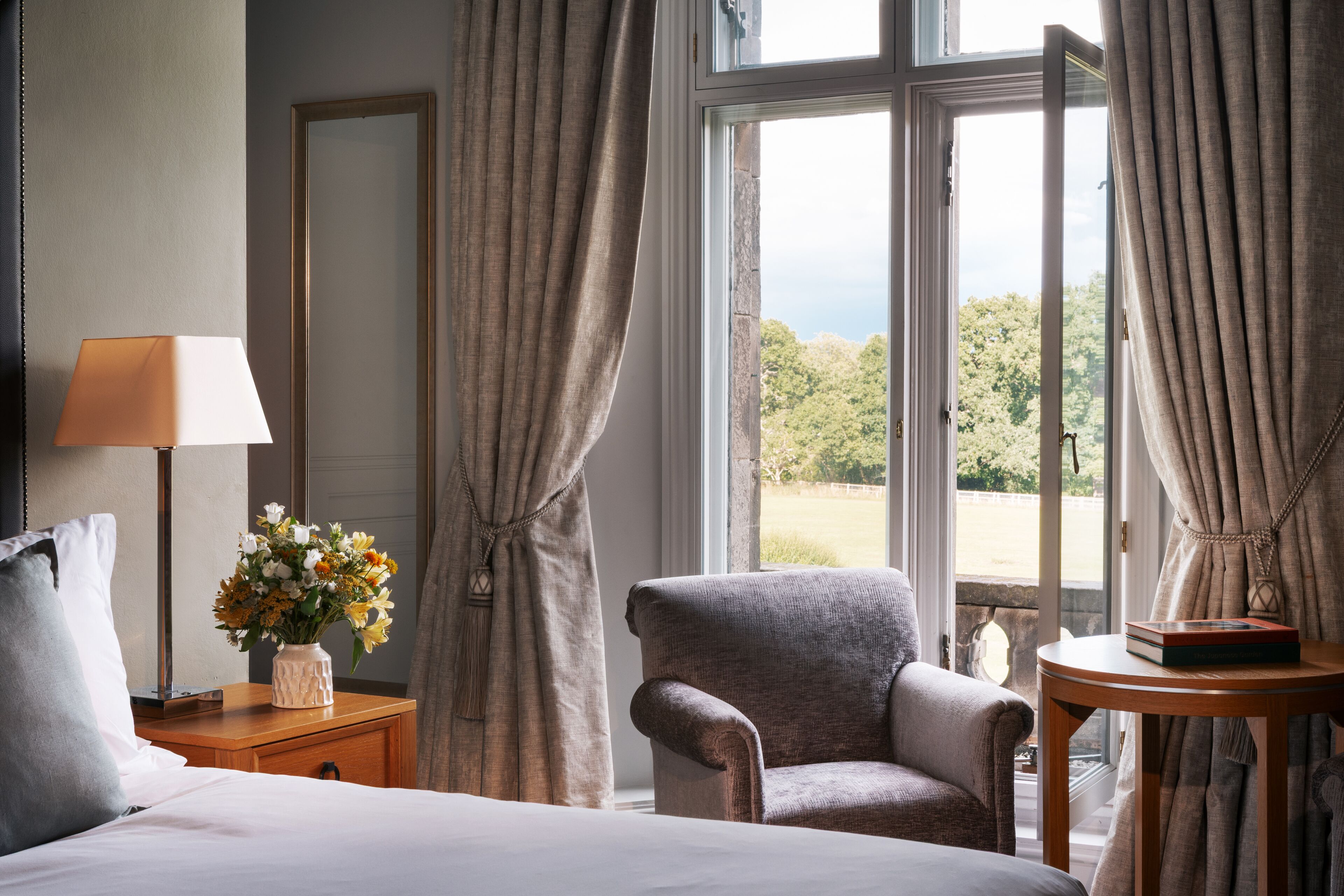 A hotel room with a bed, a chair, a nightstand with a lamp, a vase with flowers, a mirror, and a view of the outdoors through a window with curtains.