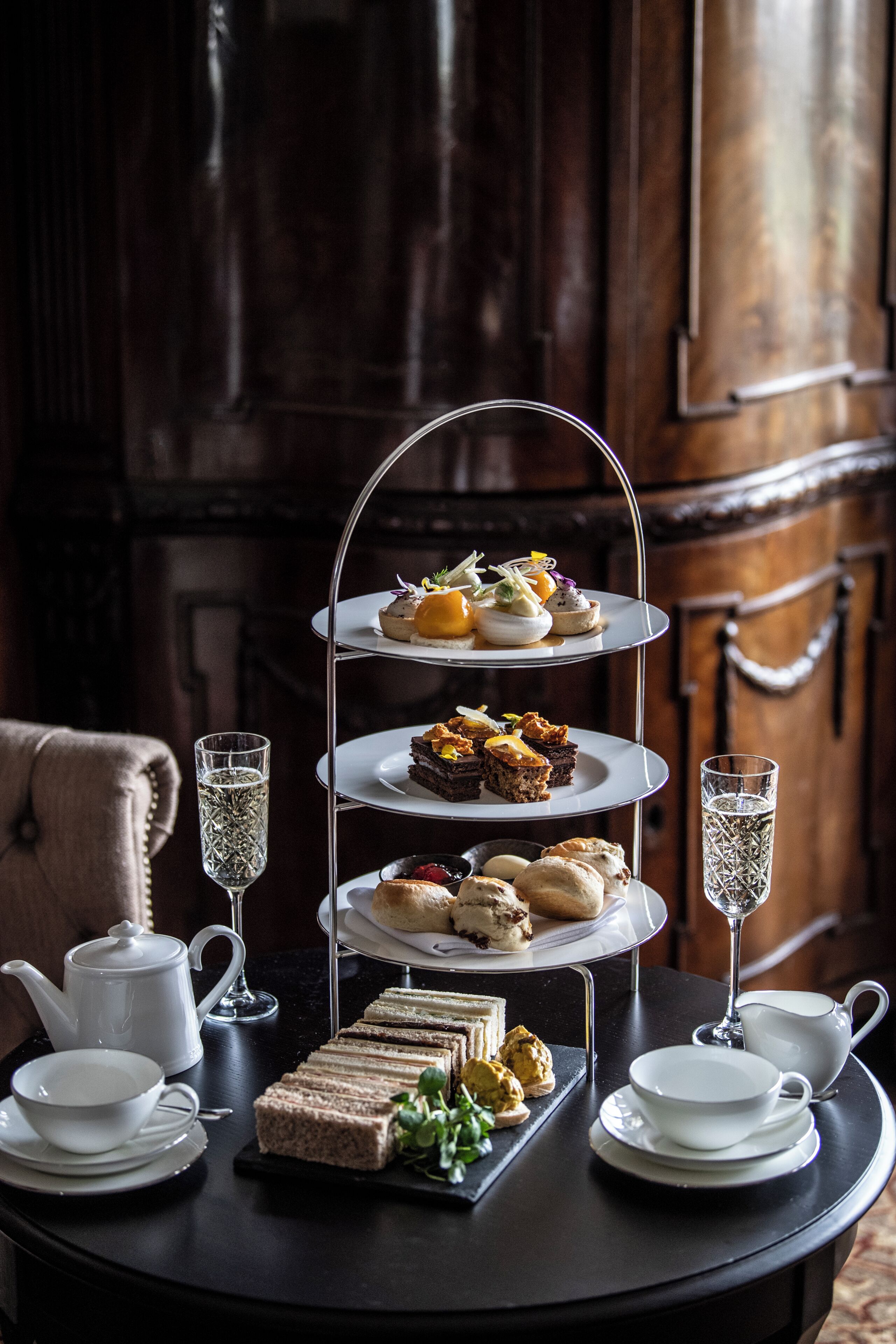 A three-tiered dessert stand with various pastries and sandwiches, accompanied by teapots, cups, and glasses on a table.