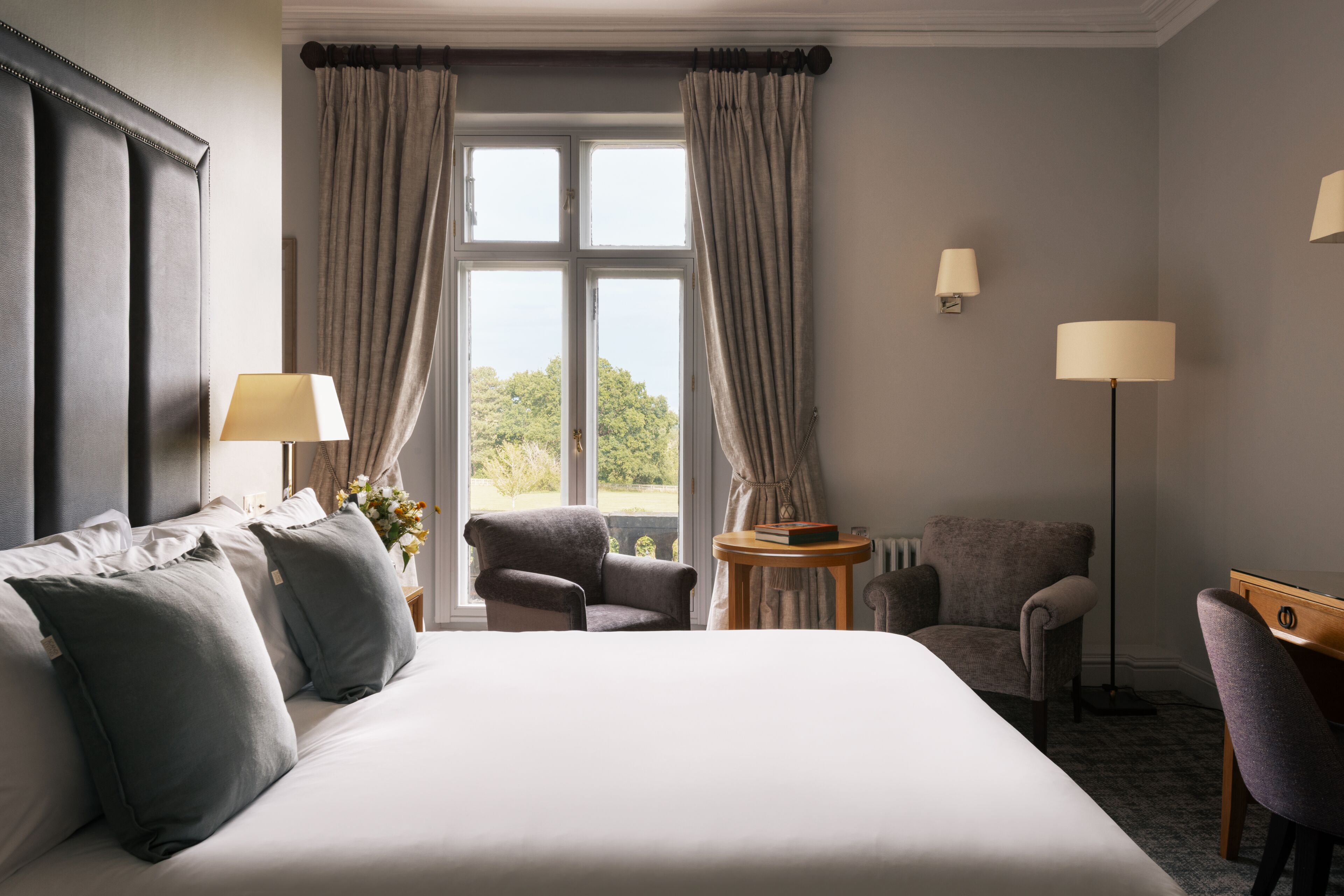 A hotel room with a large bed, two armchairs, a small table, and a window with curtains.