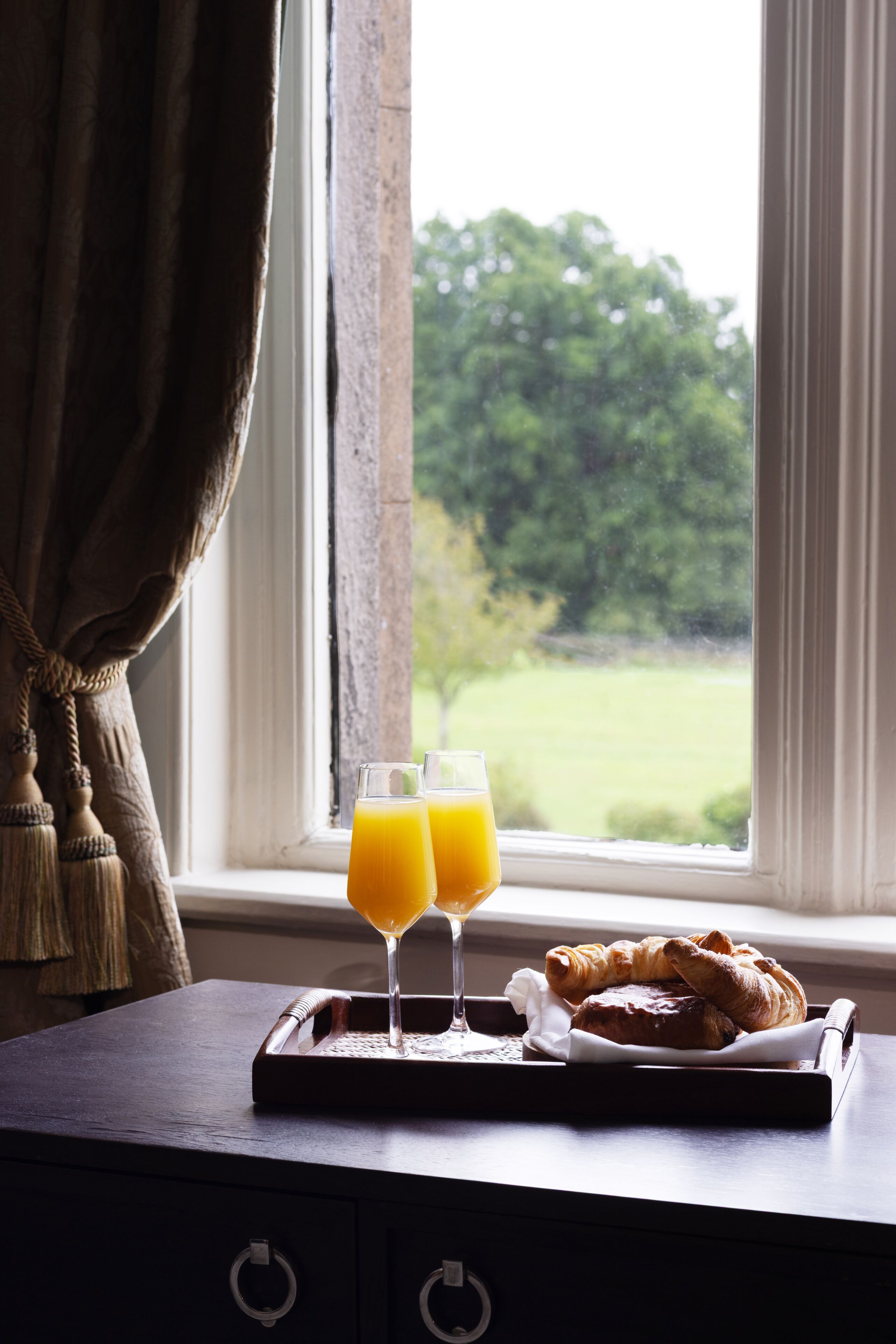 A breakfast tray with croissants and two glasses of orange juice on a table by a window with a view of greenery.