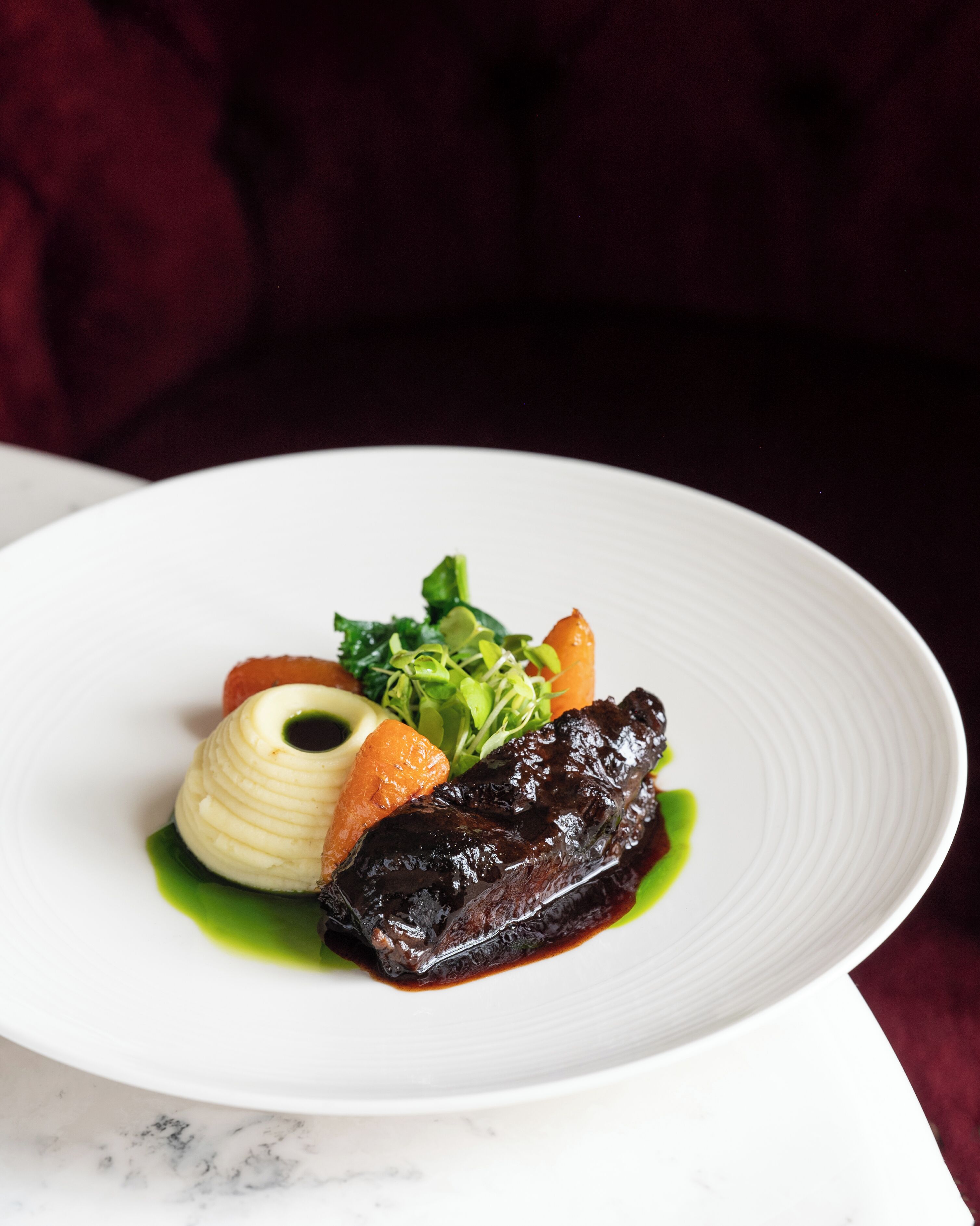 A white plate with a dish consisting of a dark meat, a carrot, and a green garnish.